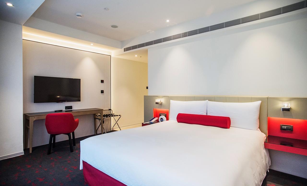 Caesar Metro Taipei Ξενοδοχείο Εξωτερικό φωτογραφία The photo shows a modern hotel room. It features a large bed with white bedding and a red accent pillow. There is a desk area with a chair, a small table, and a flat-screen television mounted on the wall. The room has a bright and minimalist design, 