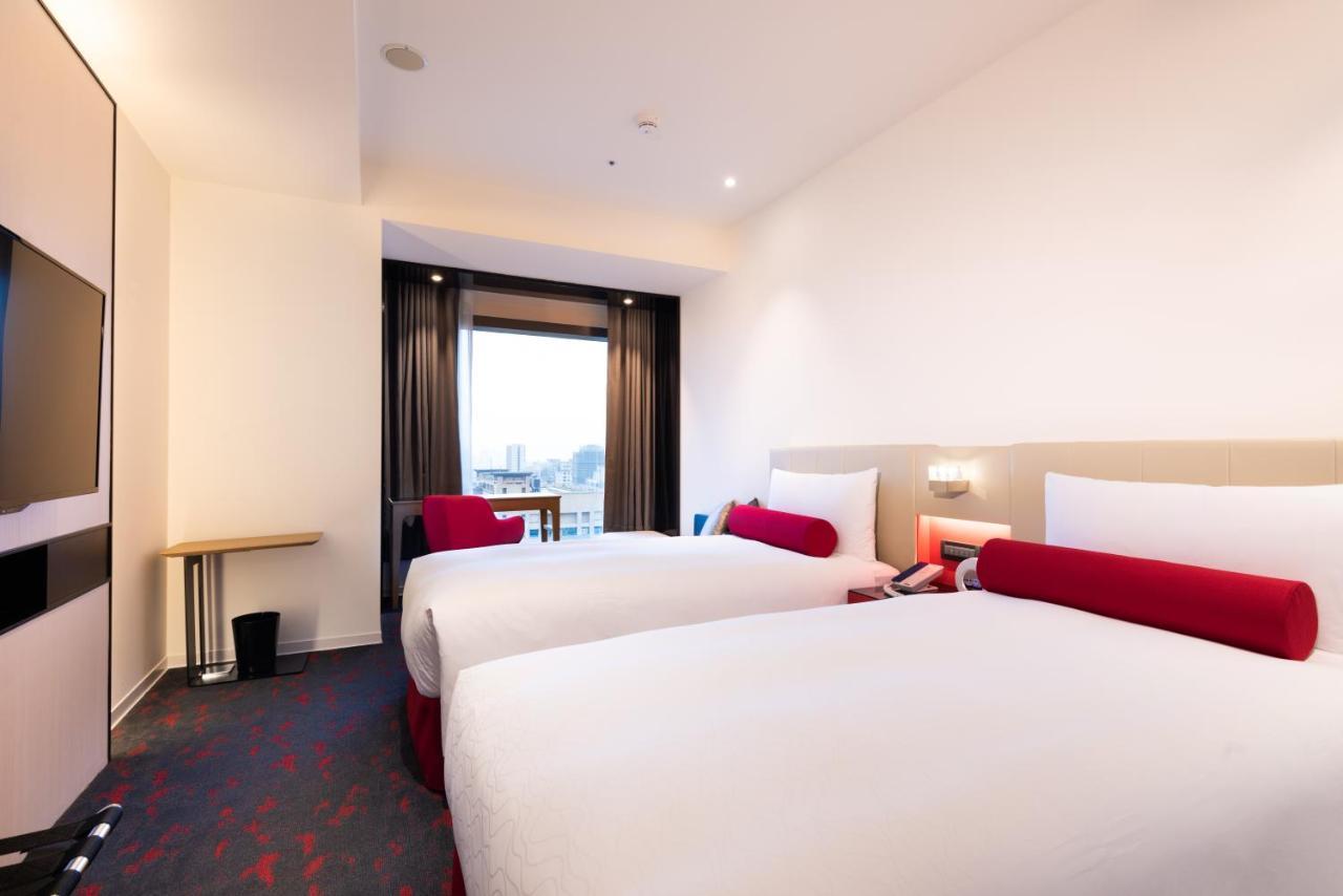 Caesar Metro Taipei Ξενοδοχείο Εξωτερικό φωτογραφία The photo shows a modern hotel room featuring two beds with white bedding and red accent pillows. There is a small wooden table next to a window that has dark curtains, allowing for natural light to enter the room. The walls are painted in light colo